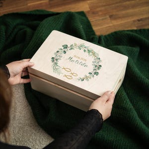 Puede incluir: Una caja de madera de color claro con tapa con bisagras. La tapa presenta un diseño de corona floral verde y dorada con el nombre "Matilda" y la fecha "15.06.2026" impresos en escritura elegante. Dos símbolos de peces dorados están debajo.