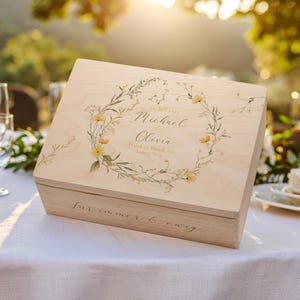 Puede incluir: Una caja de madera de color claro con un diseño de corona floral y los nombres "Michael & Olivia" impresos en la tapa. La caja está sobre un mantel blanco, con el texto "Für immer & ewig" en el frente.