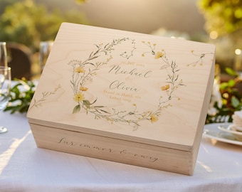 Op de afbeelding: Een lichtgekleurde houten kist met een bloemenkransontwerp en de namen "Michael & Olivia" op het deksel. De kist staat op een wit tafelkleed, met de tekst "Für immer & ewig" aan de voorkant.
