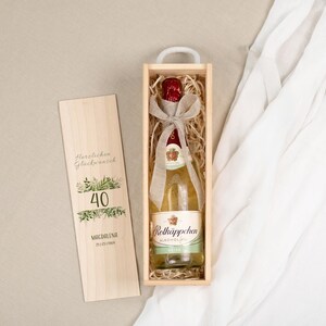 Könnte beinhalten: Eine Flasche Rotkäppchen alkoholfreier Wein in einer hölzernen Geschenkbox mit Griff. Die Flasche ist mit einem Band gebunden und liegt auf Stroh. Eine Holzkarte mit der Zahl 40 und Text liegt neben der Box.