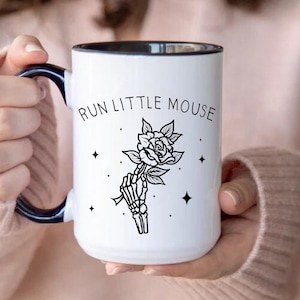 Pu&ograve; includere: Tazza in ceramica bianca con bordo e manico neri, con la scritta "RUN LITTLE MOUSE" sopra un'illustrazione in bianco e nero di una mano scheletrica che tiene una rosa. Il design include quattro piccole stelle.