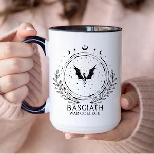 May include: White ceramic mug with a black and navy blue handle and rim. The mug features a black dragon design inside a circular frame with the text "BASGIATH WAR COLLEGE". Additional mugs with colored rims are shown.