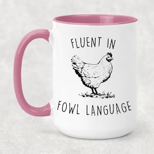 May include: White ceramic mug with a pink rim and handle. The mug has a black and white illustration of a chicken and the text "Fluent in Fowl Language".