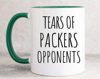 May include: White ceramic coffee mug with a green rim and handle. The mug has black text that reads "Tears of Packers Opponents".
