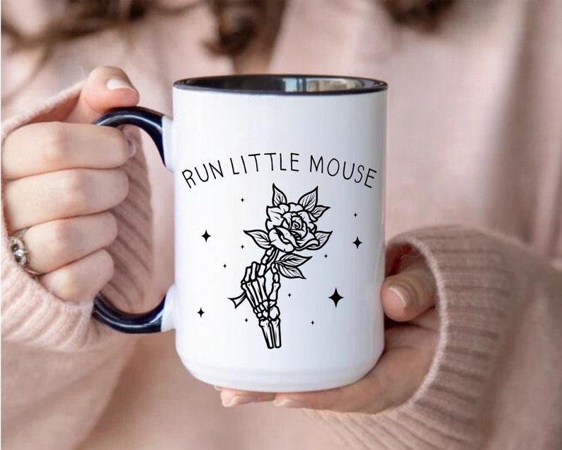 Pu&ograve; includere: Tazza in ceramica bianca con bordo e manico neri, con la scritta "RUN LITTLE MOUSE" sopra un'illustrazione in bianco e nero di una mano scheletrica che tiene una rosa. Il design include quattro piccole stelle.