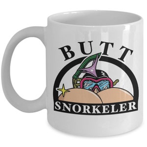 May include: White ceramic mug with a humorous design. The mug features the word "BUTT" above a cartoon image of a snorkeler with a snorkel mask and the word "SNORKELER" below. A yellow star is to the left of the snorkeler.