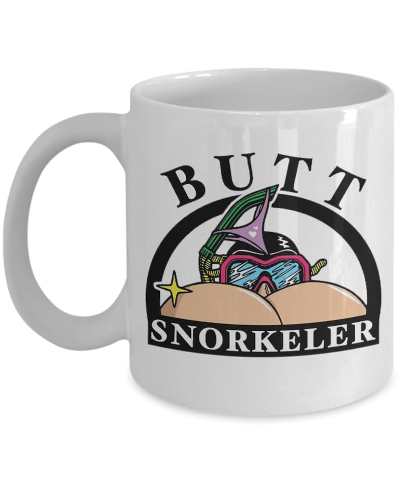 May include: White ceramic mug with a humorous design. The mug features the word "BUTT" above a cartoon image of a snorkeler with a snorkel mask and the word "SNORKELER" below. A yellow star is to the left of the snorkeler.