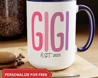 May include: White ceramic mug with a navy blue handle and interior. The mug features the name "GIGI" in pink and purple block letters, with "EST. 2023" below. A red banner at the bottom reads "PERSONALIZE FOR FREE".