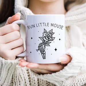 Pu&ograve; includere: Tazza in ceramica bianca con la scritta "RUN LITTLE MOUSE" e un'illustrazione in bianco e nero di una mano scheletrica che tiene una rosa. Il design include stelle. La tazza &egrave; tenuta da una persona che indossa un maglione bianco.