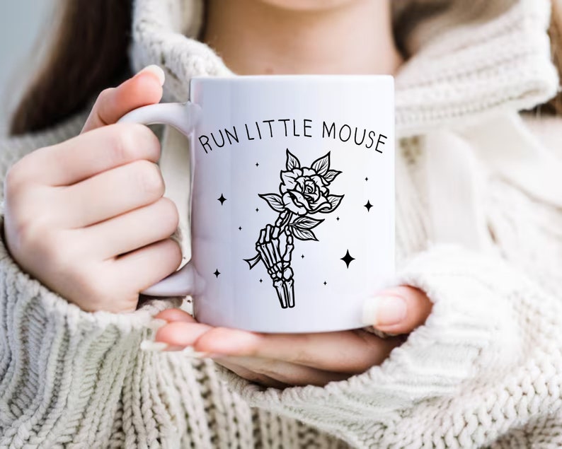 Pu&ograve; includere: Tazza in ceramica bianca con la scritta "RUN LITTLE MOUSE" e un'illustrazione in bianco e nero di una mano scheletrica che tiene una rosa. Il design include stelle. La tazza &egrave; tenuta da una persona che indossa un maglione bianco.