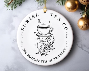 May include: White ceramic ornament with black text and a black and white illustration of a hand holding a cup of tea. The text reads "Suriel Tea Co. High Lord The Hottest Tea in Prythian Hot Tea".