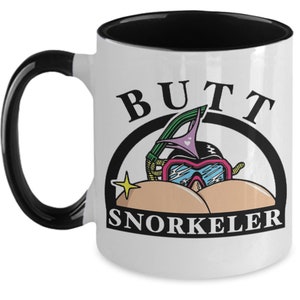 May include: White ceramic mug with a black handle and interior. The mug features a cartoon graphic of a snorkeling mask and the words "BUTT SNORKELER" in black text.