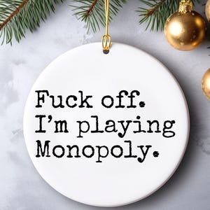 Könnte beinhalten: Weißer Keramik-Weihnachtsschmuck mit dem Text "Fuck off. I'm playing Monopoly." in Schwarz. Der Schmuck wird mit einem goldenen Faden aufgehängt, mit goldenen Kugeln und grünen Tannennadeln im Hintergrund.