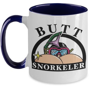 May include: White ceramic mug with a navy blue handle and interior. The mug features the text "BUTT SNORKELER" above a cartoon image of a snorkel mask and bikini bottoms. A yellow star is to the left of the text.