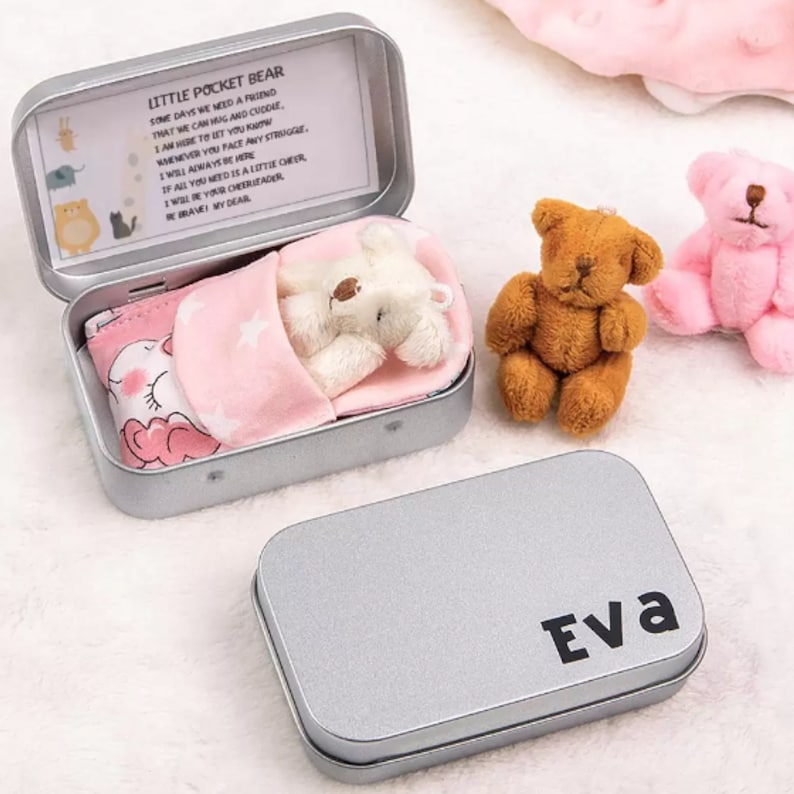 May include: A silver metal tin with a white teddy bear inside. The tin has a pink and white patterned lining and a message that reads "Little Pocket Bear. Some days we need a friend that we can hug and cuddle. I am here to let you know whenever you face any struggle. I will always be here. If all you need is a little cheer, I will be your cheerleader. Be brave, my dear." The tin is closed and has the name "Eva" printed on the top.