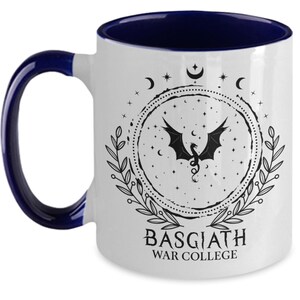 May include: White ceramic mug with a dark blue interior and handle. The mug features a black dragon design within a circular frame, surrounded by stars, moons, and leaves. The text "BASGIATH WAR COLLEGE" is printed below the design.