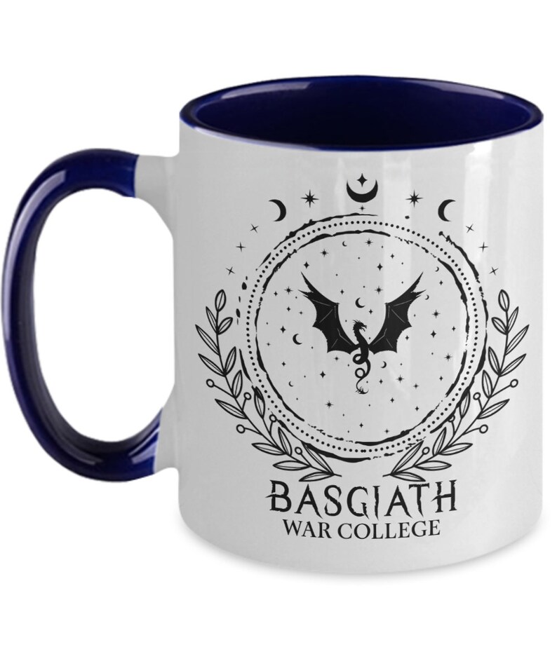May include: White ceramic mug with a dark blue interior and handle. The mug features a black dragon design within a circular frame, surrounded by stars, moons, and leaves. The text "BASGIATH WAR COLLEGE" is printed below the design.