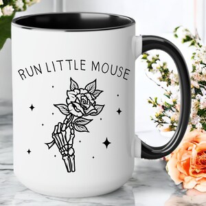 Pu&ograve; includere: Tazza in ceramica bianca con interno e manico neri. La tazza presenta la scritta "RUN LITTLE MOUSE" sopra un'illustrazione in bianco e nero di una mano scheletrica che tiene una rosa. Ci sono anche quattro stelle nere sulla tazza.