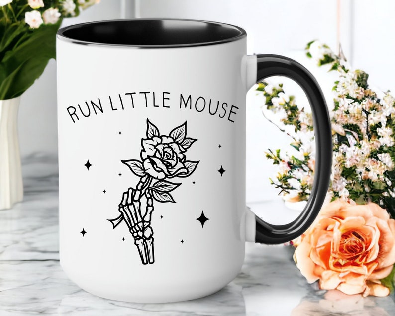 Pu&ograve; includere: Tazza in ceramica bianca con interno e manico neri. La tazza presenta la scritta "RUN LITTLE MOUSE" sopra un'illustrazione in bianco e nero di una mano scheletrica che tiene una rosa. Ci sono anche quattro stelle nere sulla tazza.