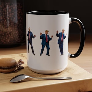 May include: White ceramic mug with a black rim and handle. The mug features a graphic of four figures in suits dancing. The figures are all wearing red ties and have blonde hair.