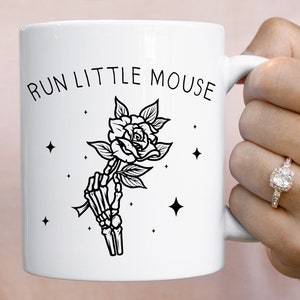 Pu&ograve; includere: Tazza in ceramica bianca con la scritta "RUN LITTLE MOUSE" e un'illustrazione nera di una mano scheletrica che tiene una rosa. Il design include stelle. La tazza &egrave; tenuta da una persona.