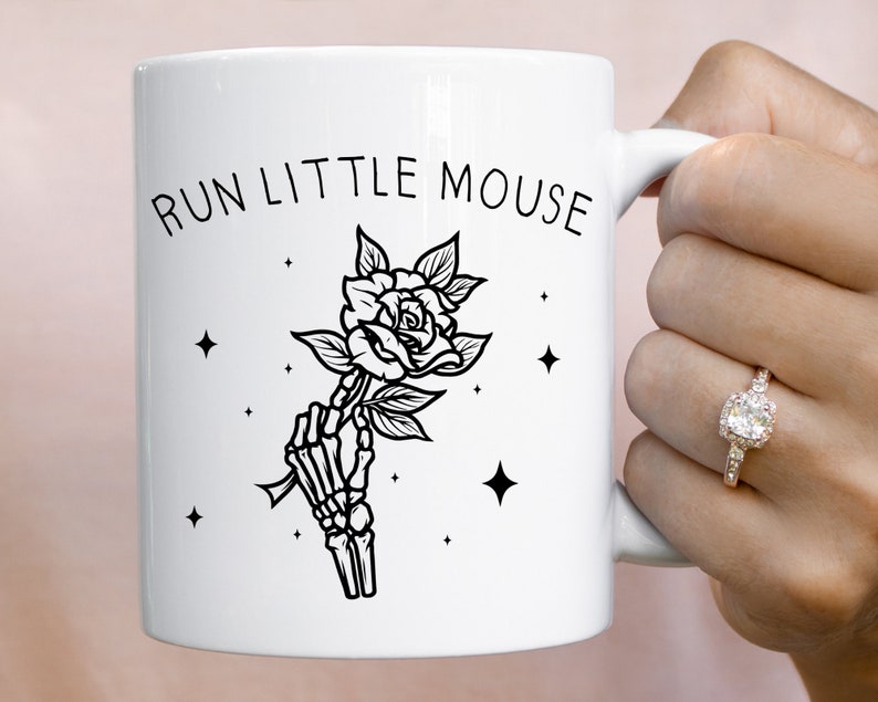 Pu&ograve; includere: Tazza in ceramica bianca con la scritta "RUN LITTLE MOUSE" e un'illustrazione nera di una mano scheletrica che tiene una rosa. Il design include stelle. La tazza &egrave; tenuta da una persona.