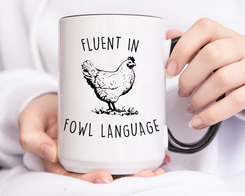 May include: White ceramic mug with a black handle and rim. The mug has a black and white illustration of a chicken and the text "Fluent in Fowl Language".