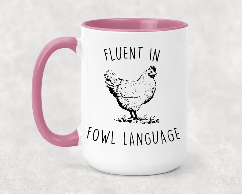 May include: White ceramic mug with a pink rim and handle. The mug has a black and white illustration of a chicken and the text "Fluent in Fowl Language".