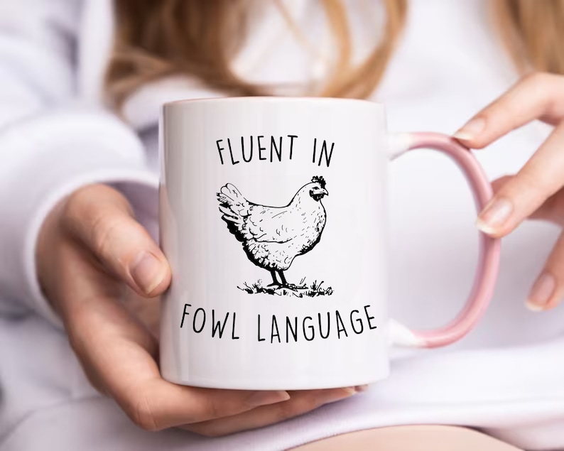 May include: White ceramic mug with a pink handle. The mug has a black and white illustration of a chicken and the text "Fluent in Fowl Language".