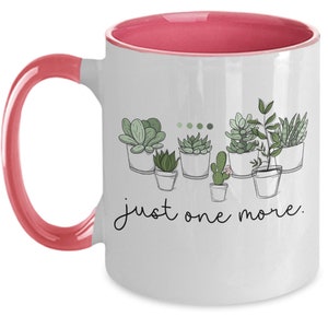 May include: White ceramic coffee mug with a pink rim and handle. The mug features a line drawing of potted succulents with the text "just one more."