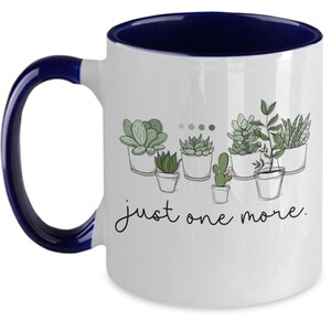 May include: White ceramic coffee mug with a blue rim and handle. The mug features a line drawing of potted succulents with the text "just one more."
