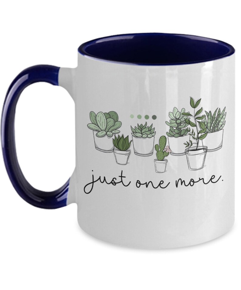 May include: White ceramic coffee mug with a blue rim and handle. The mug features a line drawing of potted succulents with the text "just one more."