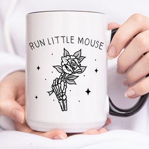 Pu&ograve; includere: Tazza in ceramica bianca con bordo e manico neri. La tazza presenta la scritta "RUN LITTLE MOUSE" sopra un'illustrazione in bianco e nero di una mano scheletrica che tiene una rosa. Sono presenti anche quattro piccole stelle.
