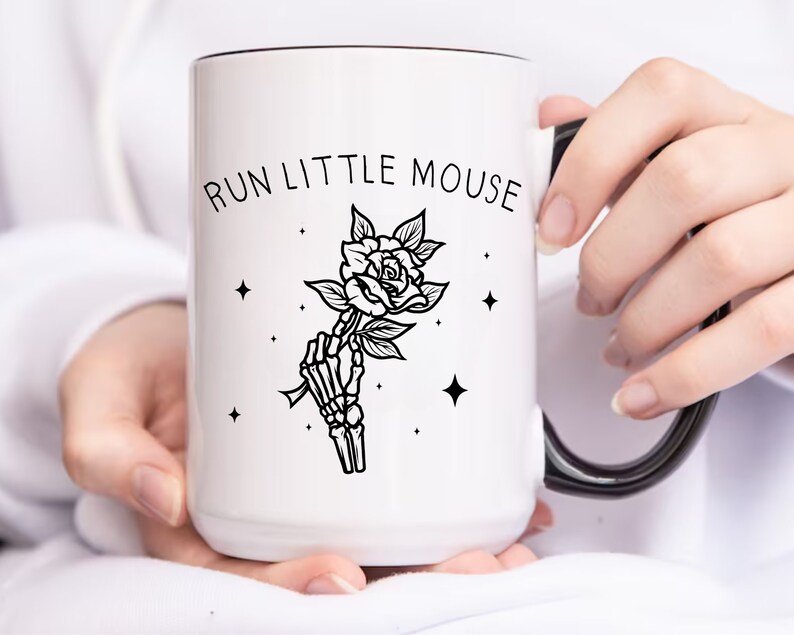 Pu&ograve; includere: Tazza in ceramica bianca con bordo e manico neri. La tazza presenta la scritta "RUN LITTLE MOUSE" sopra un'illustrazione in bianco e nero di una mano scheletrica che tiene una rosa. Sono presenti anche quattro piccole stelle.