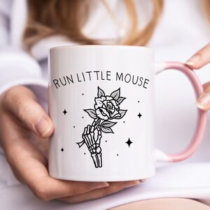 Pu&ograve; includere: Tazza in ceramica bianca con manico e bordo rosa. La tazza presenta la scritta "RUN LITTLE MOUSE" sopra un'illustrazione in bianco e nero di una mano scheletrica che tiene una rosa. Il design include piccoli accenti a stella.