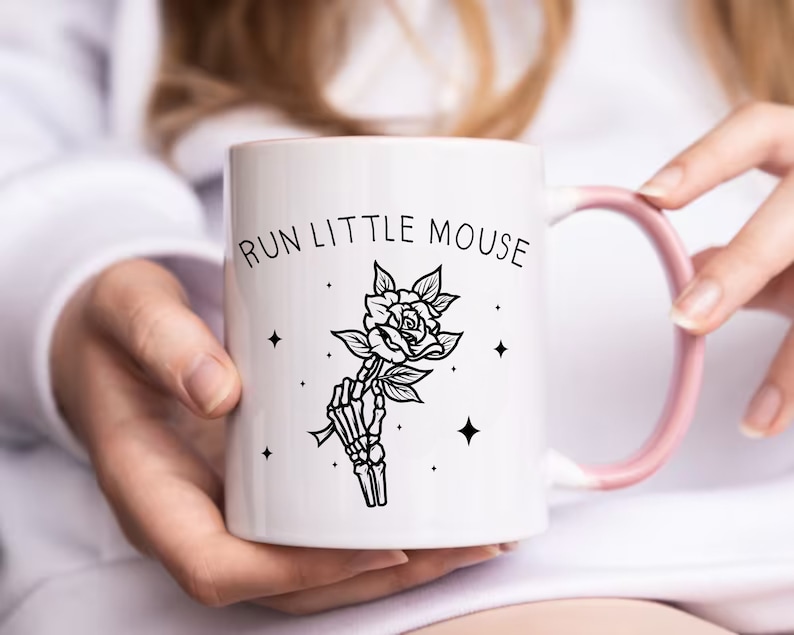 Pu&ograve; includere: Tazza in ceramica bianca con manico e bordo rosa. La tazza presenta la scritta "RUN LITTLE MOUSE" sopra un'illustrazione in bianco e nero di una mano scheletrica che tiene una rosa. Il design include piccoli accenti a stella.