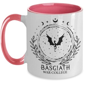 May include: White ceramic mug with a pink interior and handle. The mug features a black dragon design within a circular frame, surrounded by stars, moons, and leaves. The text "BASGIATH WAR COLLEGE" is printed below the design.