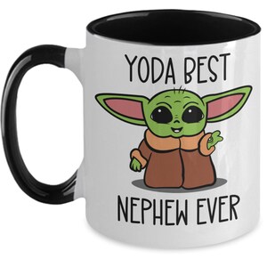 May include: White ceramic mug with a black rim and handle. The mug features a cartoon image of a green alien character with large eyes and a brown coat. The text "YODA BEST NEPHEW EVER" is printed on the mug.
