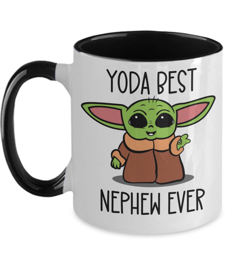 May include: White ceramic mug with a black rim and handle. The mug features a cartoon image of a green alien character with large eyes and a brown coat. The text "YODA BEST NEPHEW EVER" is printed on the mug.