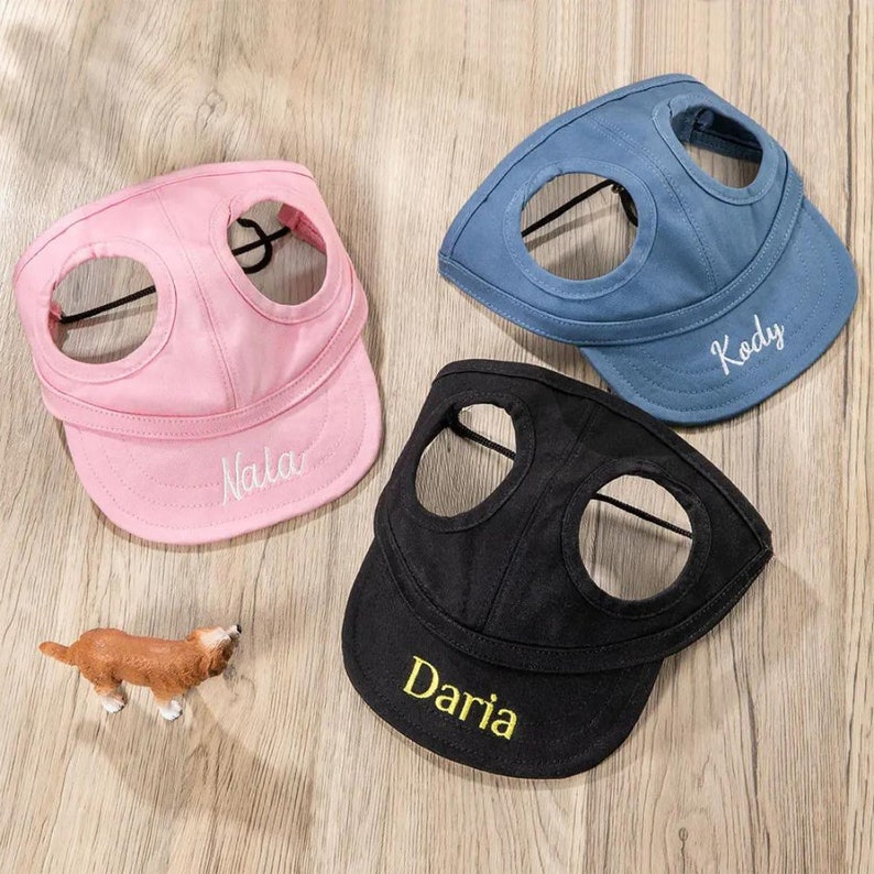 May include: Three dog hats in pink, blue, and black with the names "Nala", "Kody", and "Daria" embroidered on them. The hats have two holes for the dog's ears and a strap to secure the hat.
