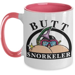 May include: White ceramic mug with a pink interior and handle. The mug features the words "BUTT SNORKELER" in black, with a cartoon illustration of a snorkel mask and goggles. A yellow star is also included in the design.