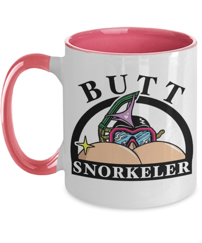 May include: White ceramic mug with a pink interior and handle. The mug features the words "BUTT SNORKELER" in black, with a cartoon illustration of a snorkel mask and goggles. A yellow star is also included in the design.