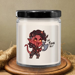 May include: A glass candle jar with a black lid. The jar features a cartoon illustration of a red demon holding an axe, with the text "OH FUCK YES!". The candle sits on a wooden surface.