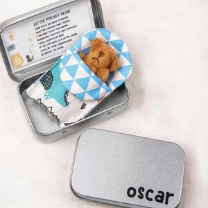 May include: A small, brown teddy bear wearing a blue and white patterned sleeping bag is inside a silver tin with the words "Little Pocket Bear" and a message about friendship. The tin has a silver lid with the name "oscar" printed on it.