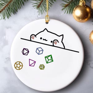 May include: Round white ceramic Christmas ornament with a cartoon cat design peeking over a black line, and colourful dice illustrations below. The ornament has a gold string for hanging. Gold baubles and evergreen branches are in the background.