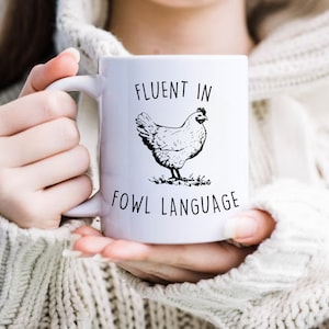 May include: White ceramic mug with a black and white illustration of a chicken and the text "Fluent in Fowl Language".