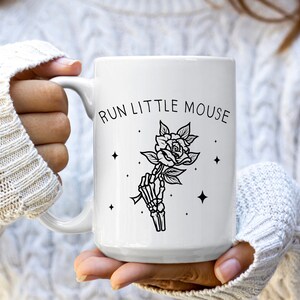 Pu&ograve; includere: Tazza in ceramica bianca con una grafica nera di una mano di scheletro che tiene una rosa, con la scritta "RUN LITTLE MOUSE" sopra. La tazza ha un manico grande ed &egrave; tenuta da una persona.