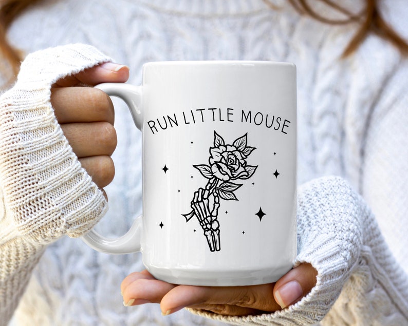 Pu&ograve; includere: Tazza in ceramica bianca con una grafica nera di una mano di scheletro che tiene una rosa, con la scritta "RUN LITTLE MOUSE" sopra. La tazza ha un manico grande ed &egrave; tenuta da una persona.