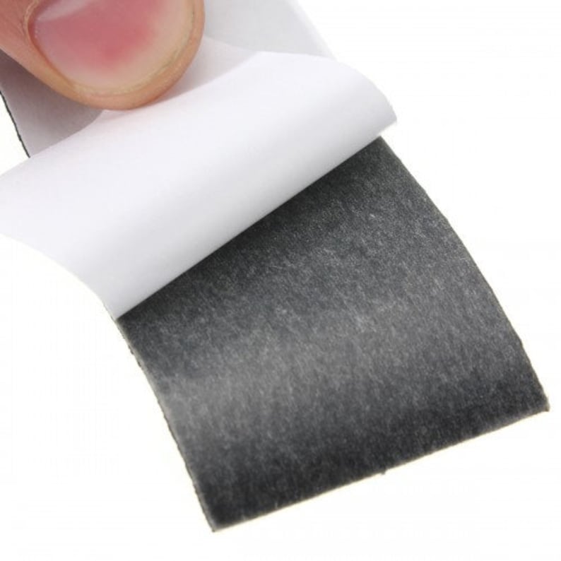 May include: Close-up of a square, dark gray adhesive pad with a white backing being peeled away. The pad has a textured, felt-like appearance. The background is white.