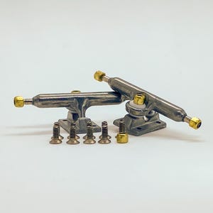 Pode incluir: Dois trucks de skate prateados com hardware dourado e um conjunto de parafusos correspondentes. Os trucks estão angulados, mostrando seu design e componentes. O hardware inclui parafusos e porcas, prontos para montagem.